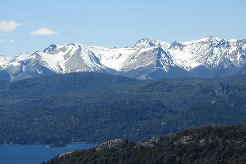 Vista de las montañas nevadas de la cordillera y el bosque andino desde el cerro Campanario, con el lago Nahuel Huapi asomando abajo, cerca de Bariloche