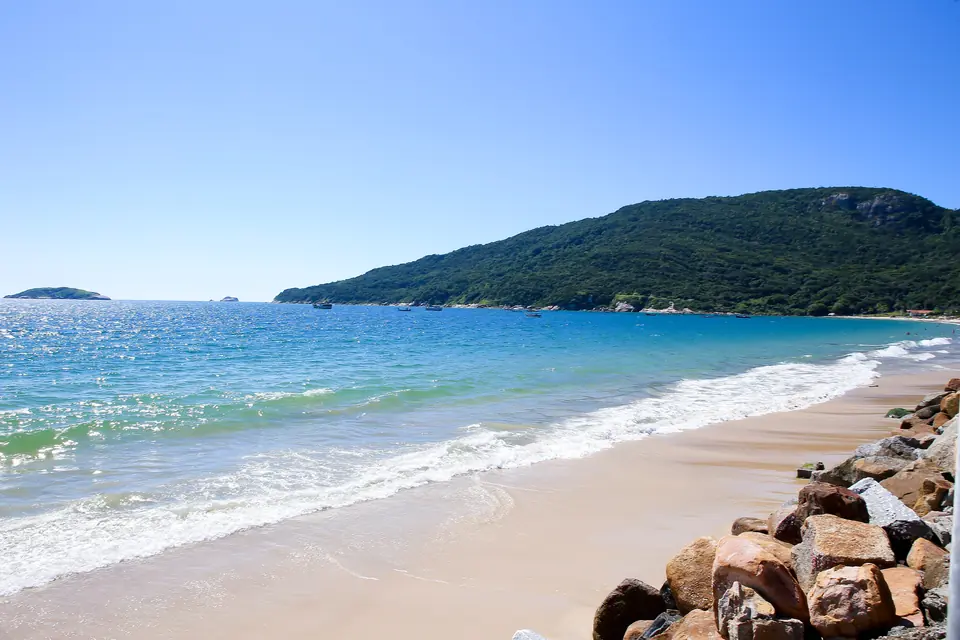 Una playa vacía y tranquila de Florianópolis, con agua turquesa en degradé, arena clara y un cerro verde al fondo
