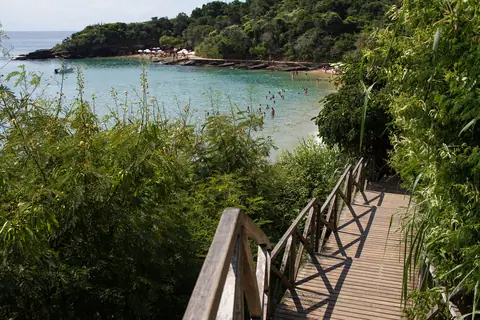 Praia Azeda, en Búzios: una pasarela de madera baja entre la vegetación hacia una playa chica de agua turquesa, con un morro verde al fondo