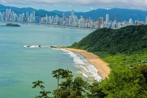 Vista aérea de la Praia do Buraco: mar verde, un morro con selva y, al fondo, la línea de edificios de Balneário Camboriú frente a la bahía