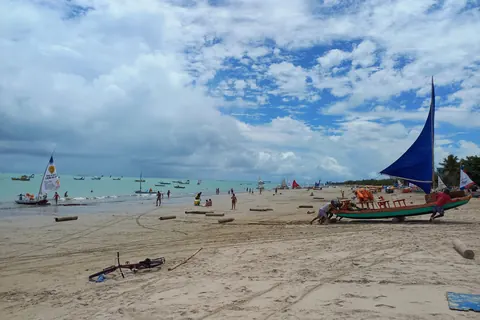 Praia de Pajuçara, en Maceió: una jangada con vela azul sobre la arena, el mar verde claro y otras velas a lo lejos bajo un cielo con nubes