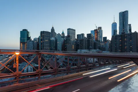 Los rascacielos del Lower Manhattan al atardecer vistos desde el puente de Brooklyn, con las estelas de luz de los autos sobre el tablero del puente