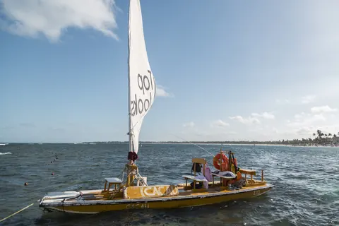 Una jangada de vela blanca navega hacia las piscinas naturales de Porto de Galinhas, con el mar verde y la costa de cocoteros al fondo