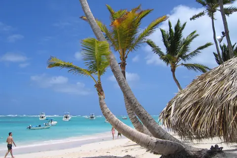 Playa Bávaro, en Punta Cana: cocoteros inclinados sobre la arena blanca, un techo de palma a la derecha y lanchas en el mar turquesa